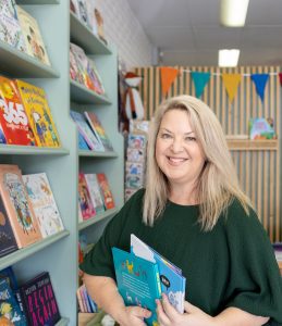 A woman with blonde hair holds picture books. She is standing in a bookshop and smiling. 