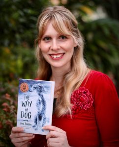 A woman is smiling. She wears a red top. She is holding a book titled The Way Of Dog. 